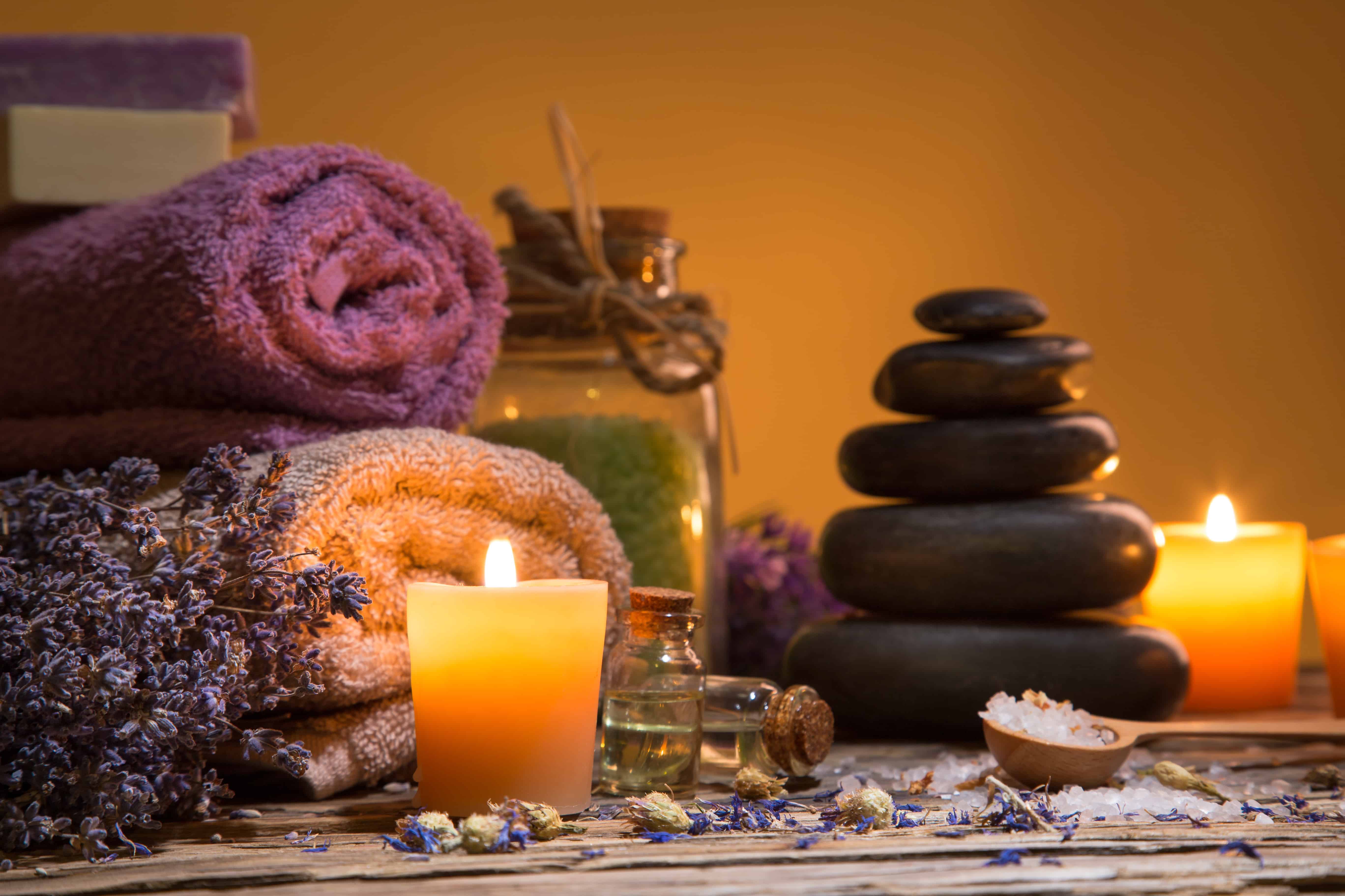 Image of spa items like a candle, salt, oil, lavendar, towels, stones, soap, and jars on a table. 