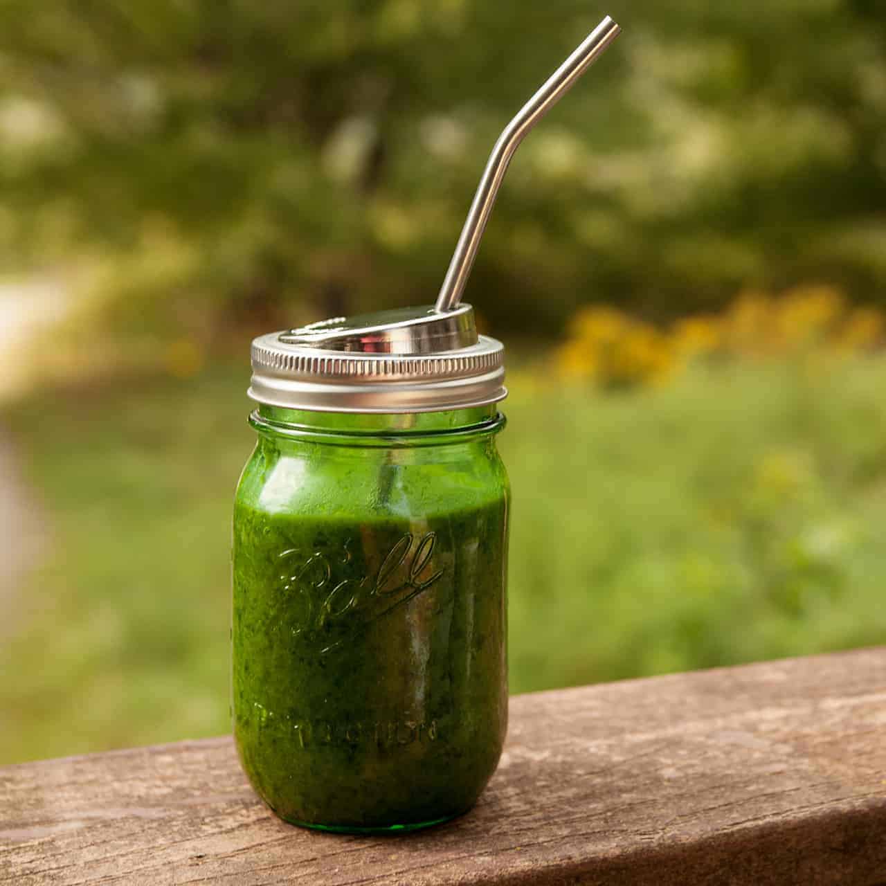 A jar with a straw and green juice on a wooden platform with trees and grass in the background