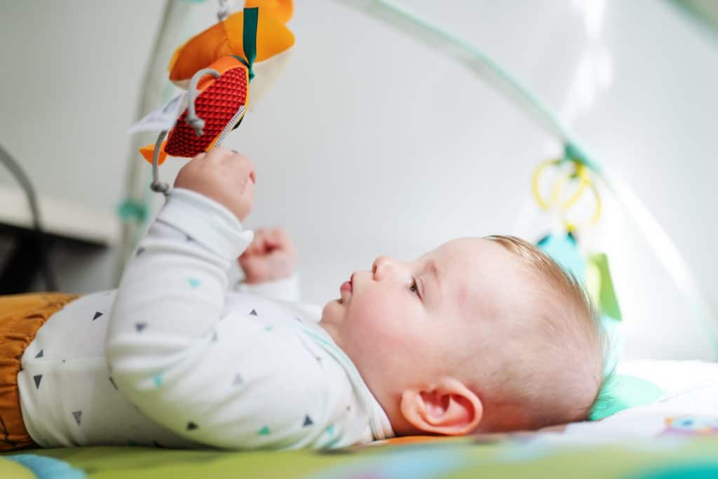Side view of beautiful curious caucasian six months old baby boy lying in bed and playing with crib non-toxic wood baby gym toys.