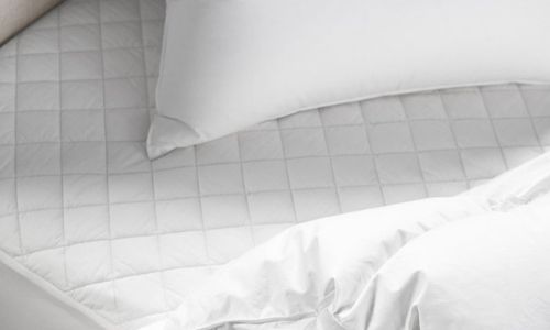 Coyuchi organic cotton mattress protector shown on a mattress with a pillow and comforter, all in white. 