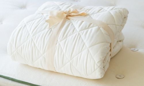 white GOTS certified organic cotton mattress protector folded on a bed with a beige ribbon around it