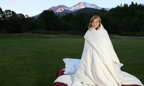 girl standing in grass field with mountains and trees in the background.  She is wrapped up in an organic comforter. 