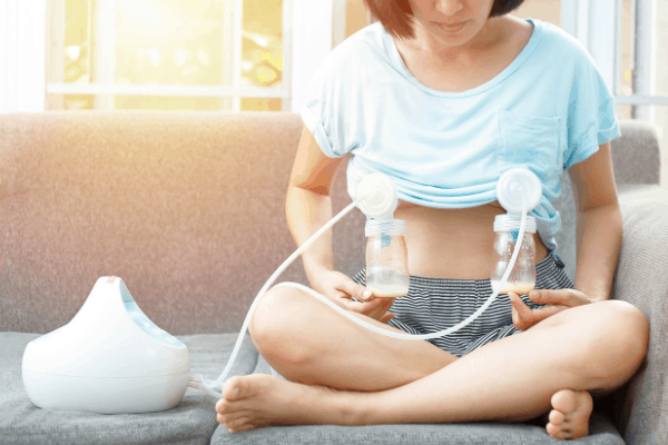 woman sitting on couch in blue shirt and dark blue shorts pumping breast milk with her non-toxic breast pump